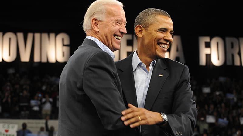 Joe Biden y Barack Obama ambos en trajes oscuros, abrazándose y sonriendo.